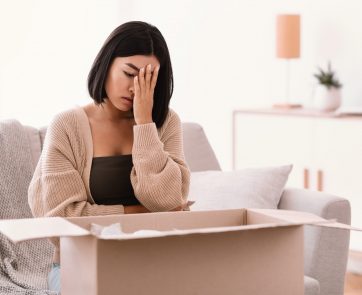 Wrong Parcel. Unhappy frustrated asian woman unpacking box sad with bad delivery service sitting on sofa at home in living room. Shipping mistake, copy space. Lady covering face with palm